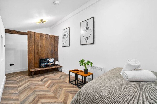 A modern bedroom with a herringbone floor, a wooden bench, a TV, a small table with a plant, and a bed with a rolled towel.