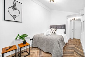 A bedroom with a grey upholstered bed, a potted plant, a wooden side table, a framed artwork, and a herringbone-patterned floor.