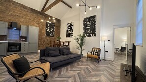 Ein modernes Wohnzimmer mit Fischgrätparkett, einem grauen Sofa, einem Korbstuhl und einem Essbereich mit Backsteinwand und Holzbalkendecke.