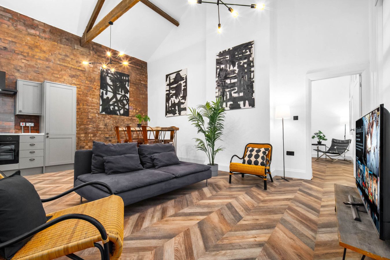 Una sala moderna con un sofá gris, un sillón amarillo y un comedor con mesa y sillas de madera. Se observan obras de arte abstracto en la pared, de color negro, y una planta en maceta.
