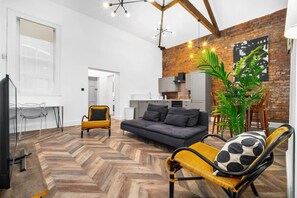 A modern living room with a herringbone floor, a grey sofa, and a yellow armchair. There is a television, a dining table with chairs, and a kitchen area visible in the background. - Stylish 2 Bedroom Apartment - Netflix - Wolverhampton Centre (Wolverhampton)