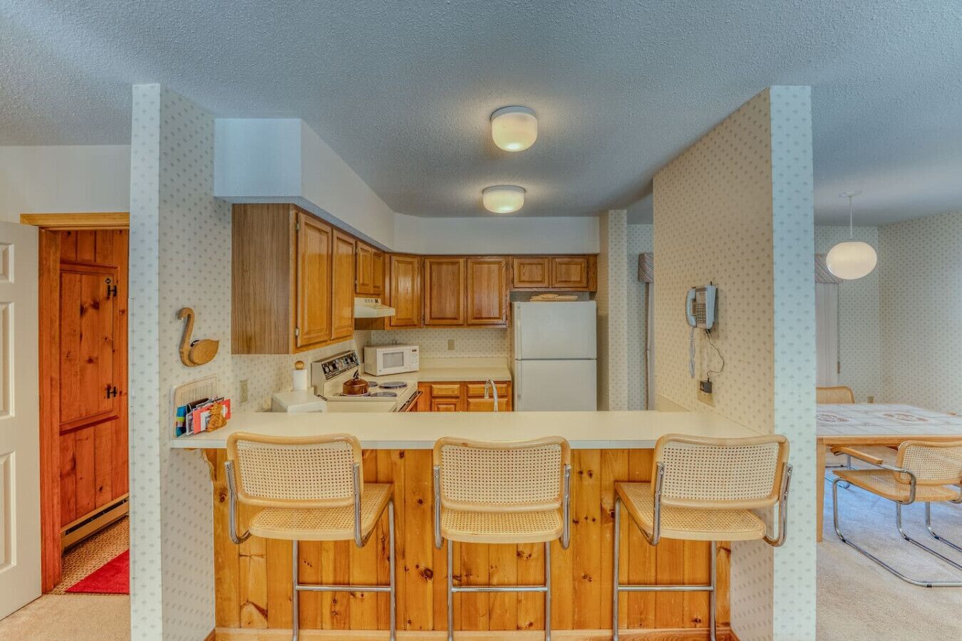 A compact kitchen with wooden cabinets, a microwave, and a refrigerator. There are three wicker bar stools at the counter.