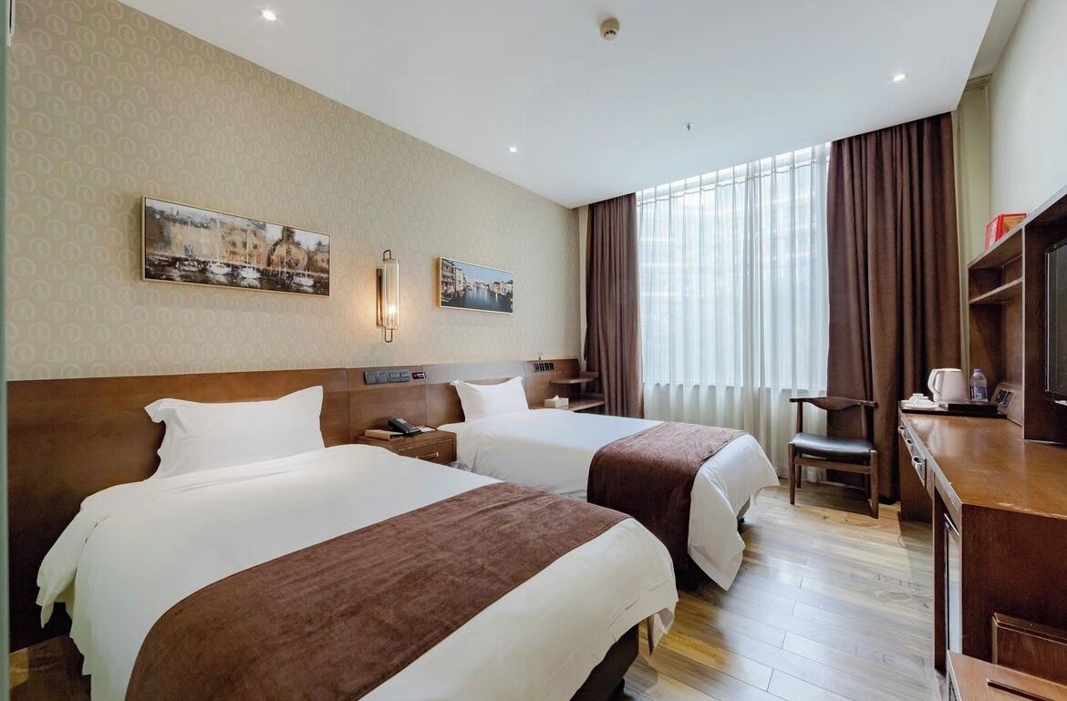 A hotel room with two beds, a desk, a chair, a TV, and a window with curtains.