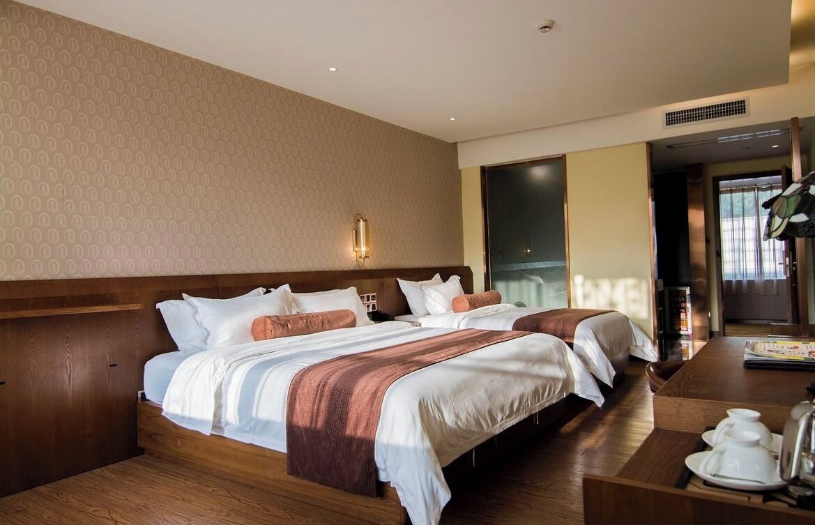 A hotel room with two beds, a wooden headboard, a bedside table with a kettle, and a large window.