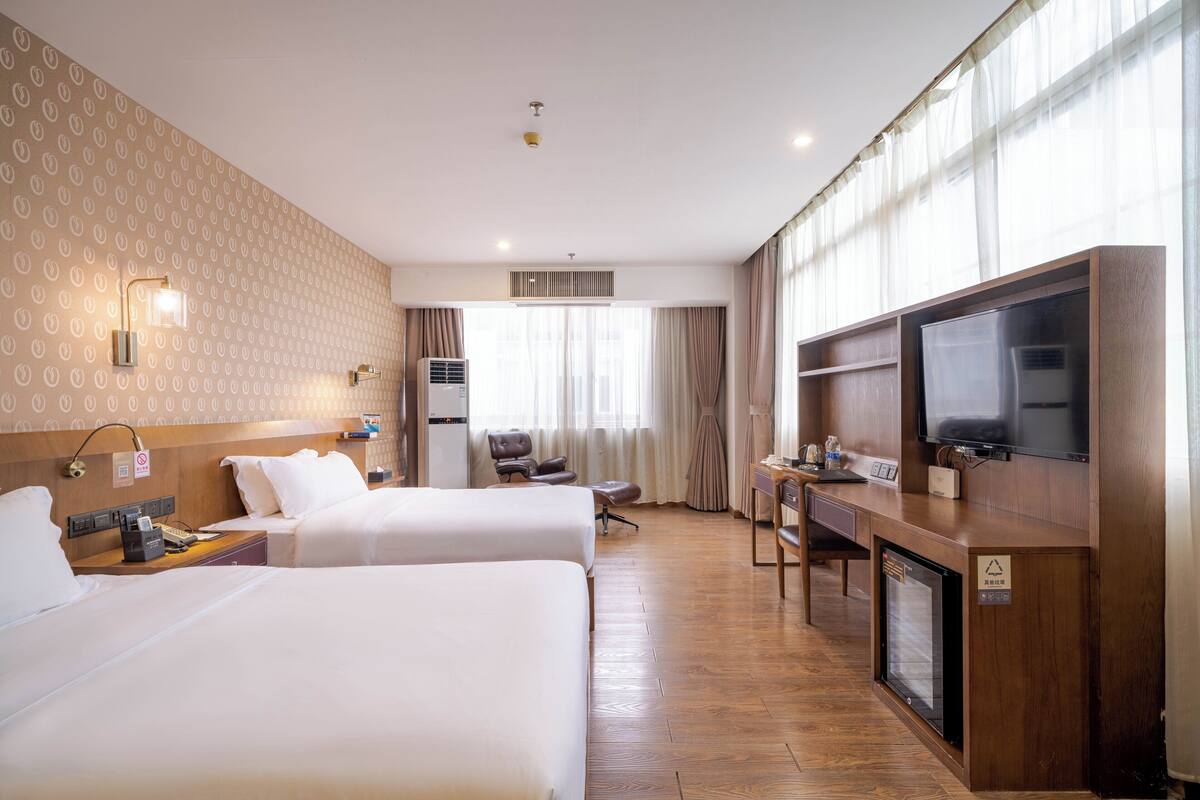 Une chambre d’hôtel avec deux lits, un bureau avec une télévision, un minibar et une grande fenêtre avec des rideaux.