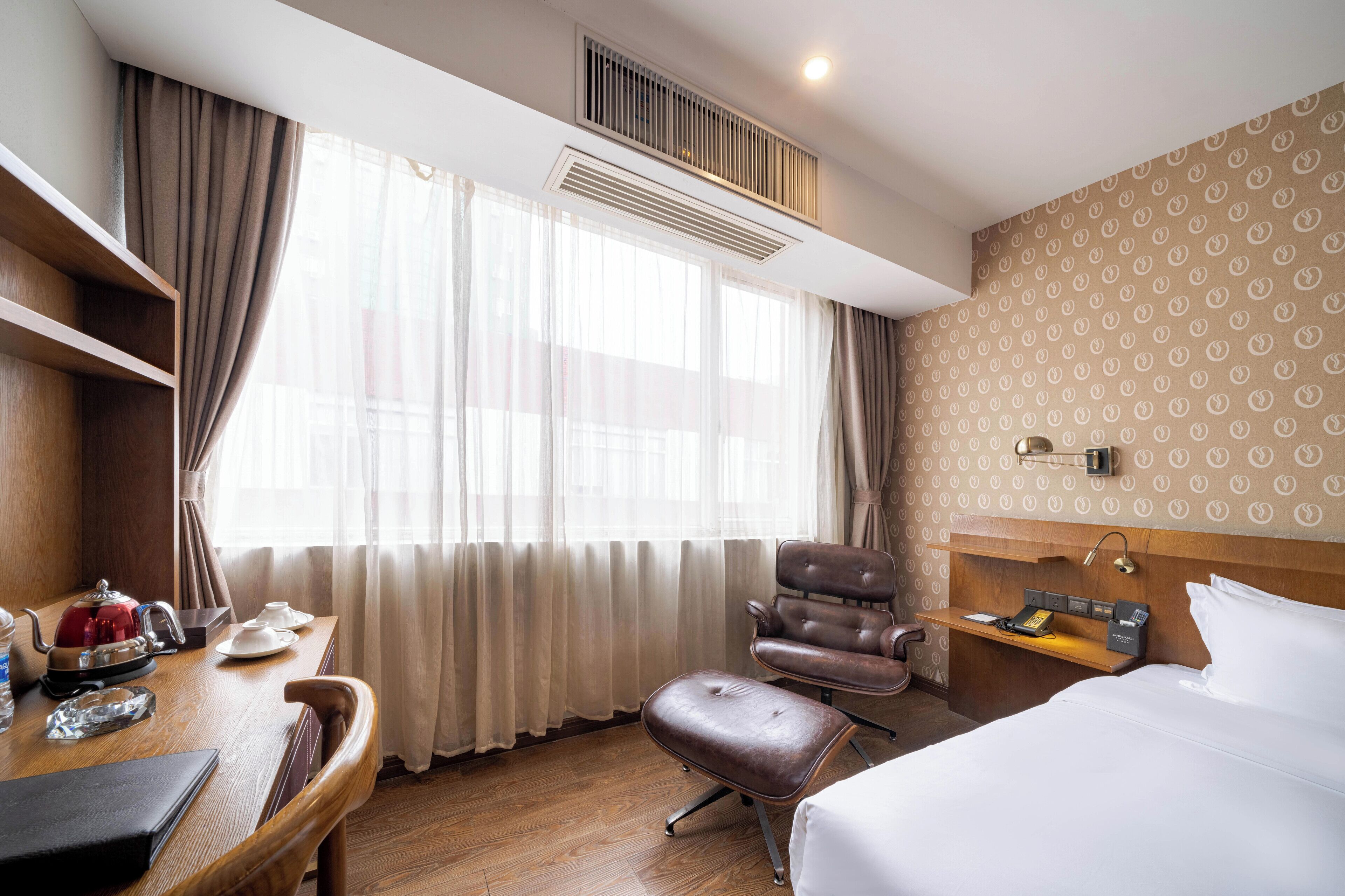 A hotel room with a bed, a desk with a kettle, a chair, and a bedside table with a lamp.