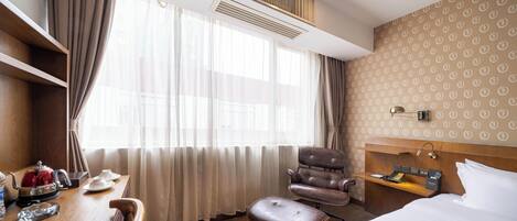 A hotel room with a bed, a desk with a kettle, a chair, and a bedside table with a lamp.