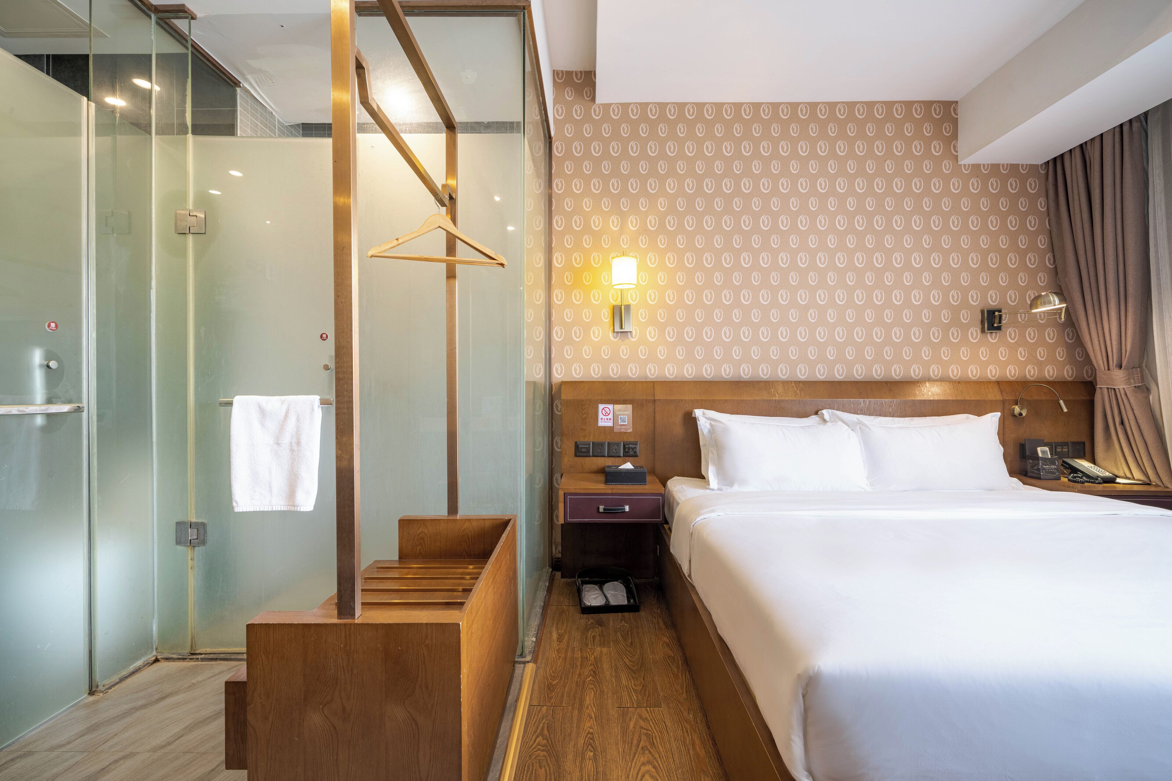A hotel room with a large bed, a wooden bench, a glass shower enclosure, and a wall-mounted light fixture.