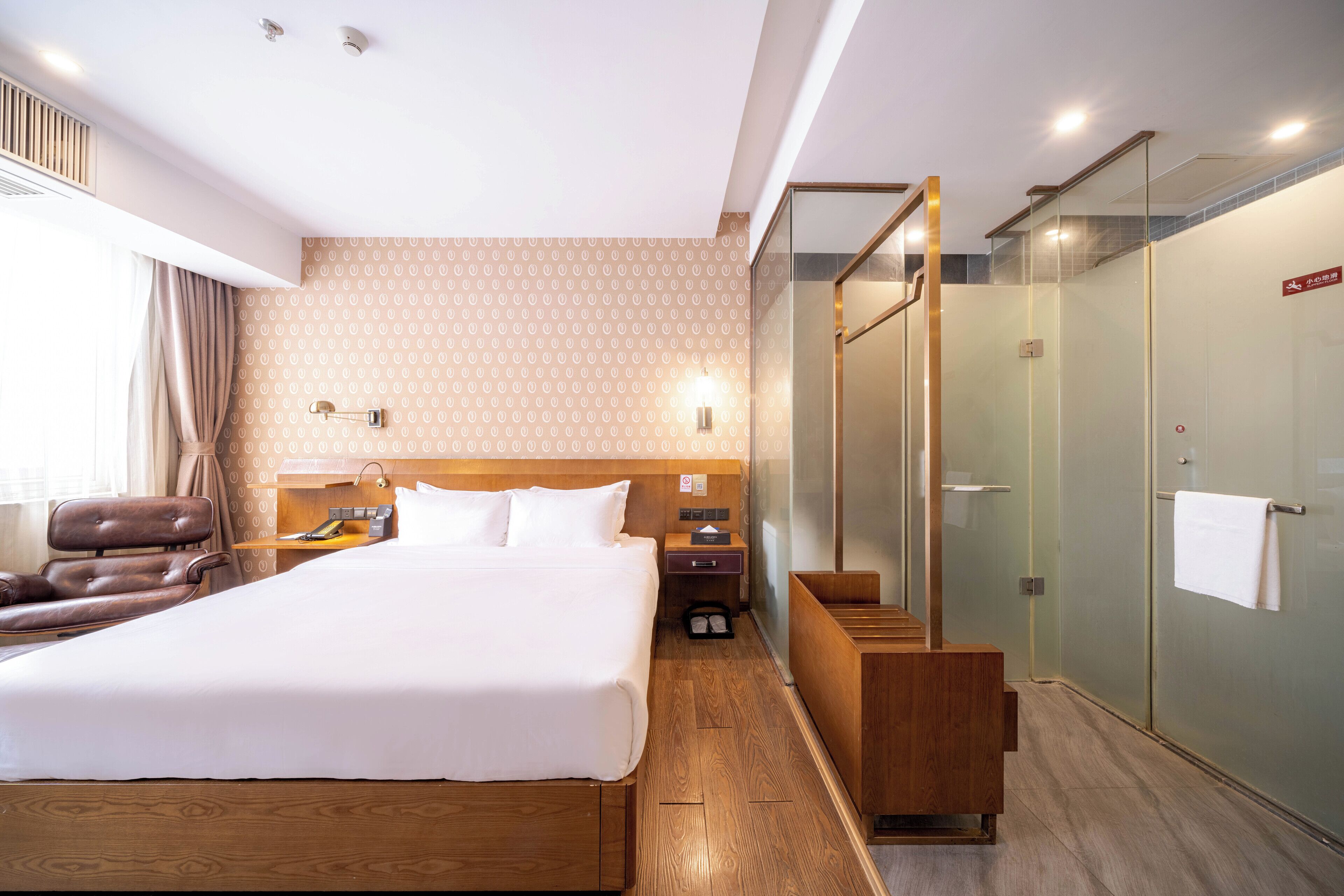 A hotel room with a large bed, a wooden headboard, a desk with a chair, a leather armchair, a glass-enclosed shower, and a wooden bench.