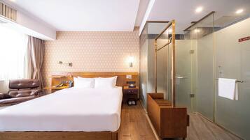 A hotel room with a large bed, a wooden headboard, a desk with a chair, a leather armchair, a glass-enclosed shower, and a wooden bench.