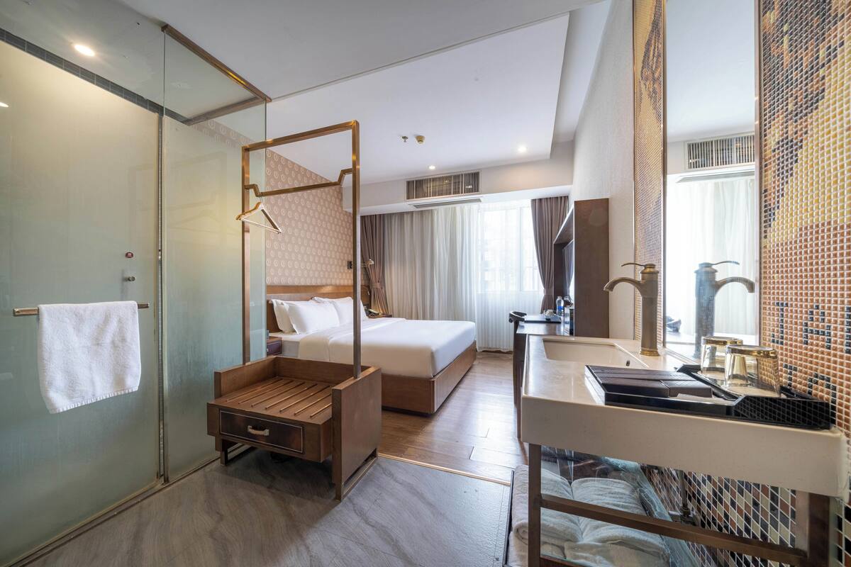 Une chambre d’hôtel dotée d’une douche en verre, d’un cadre de lit en bois, d’une table de chevet avec une lampe et d’une grande fenêtre avec des rideaux.
