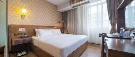 Habitación de hotel con una cama grande, un escritorio con silla, una mesita de noche, una lámpara de pared y una ventana con cortinas.