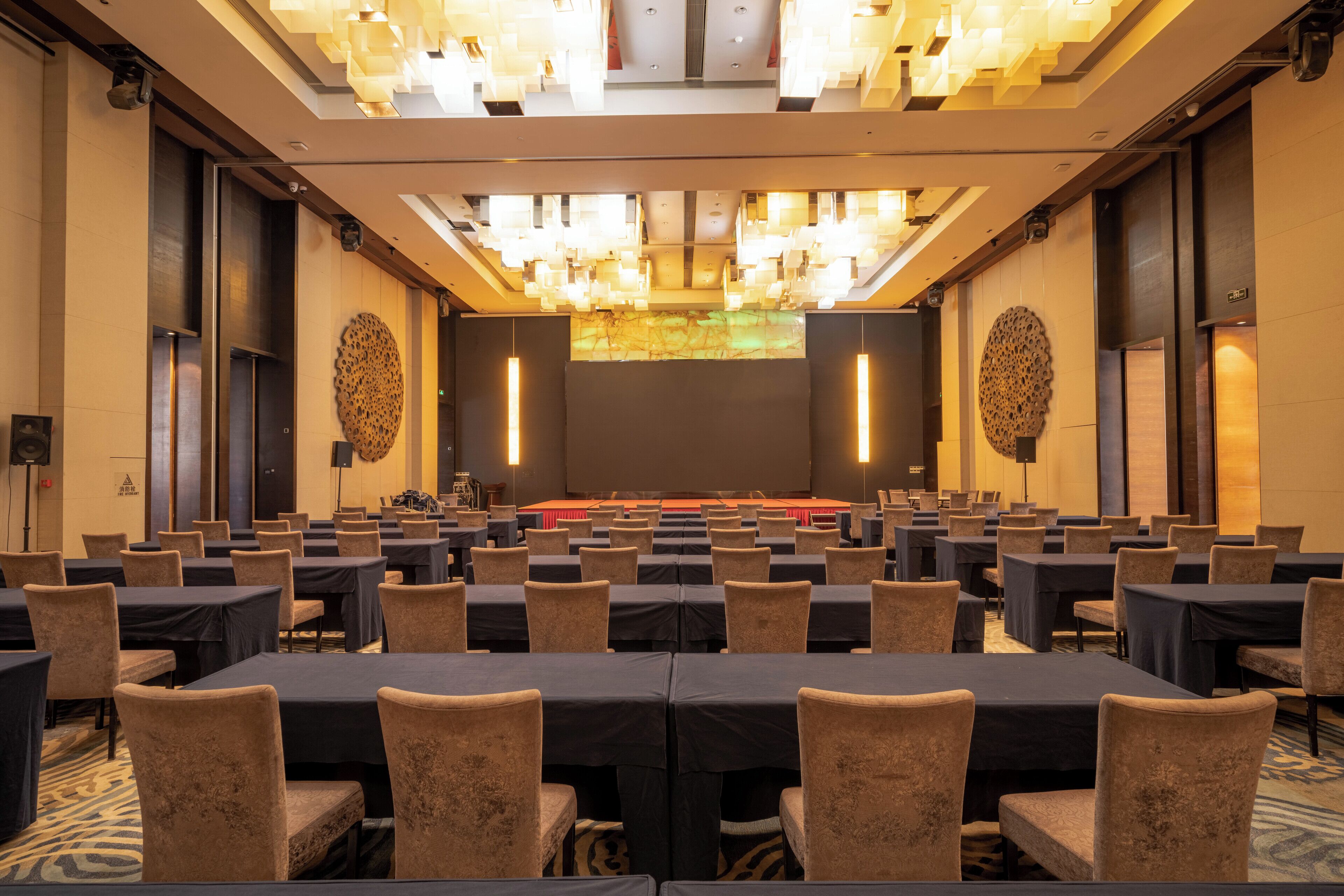 A conference room with rows of tables and chairs, a stage with a red carpet, and a large screen.
