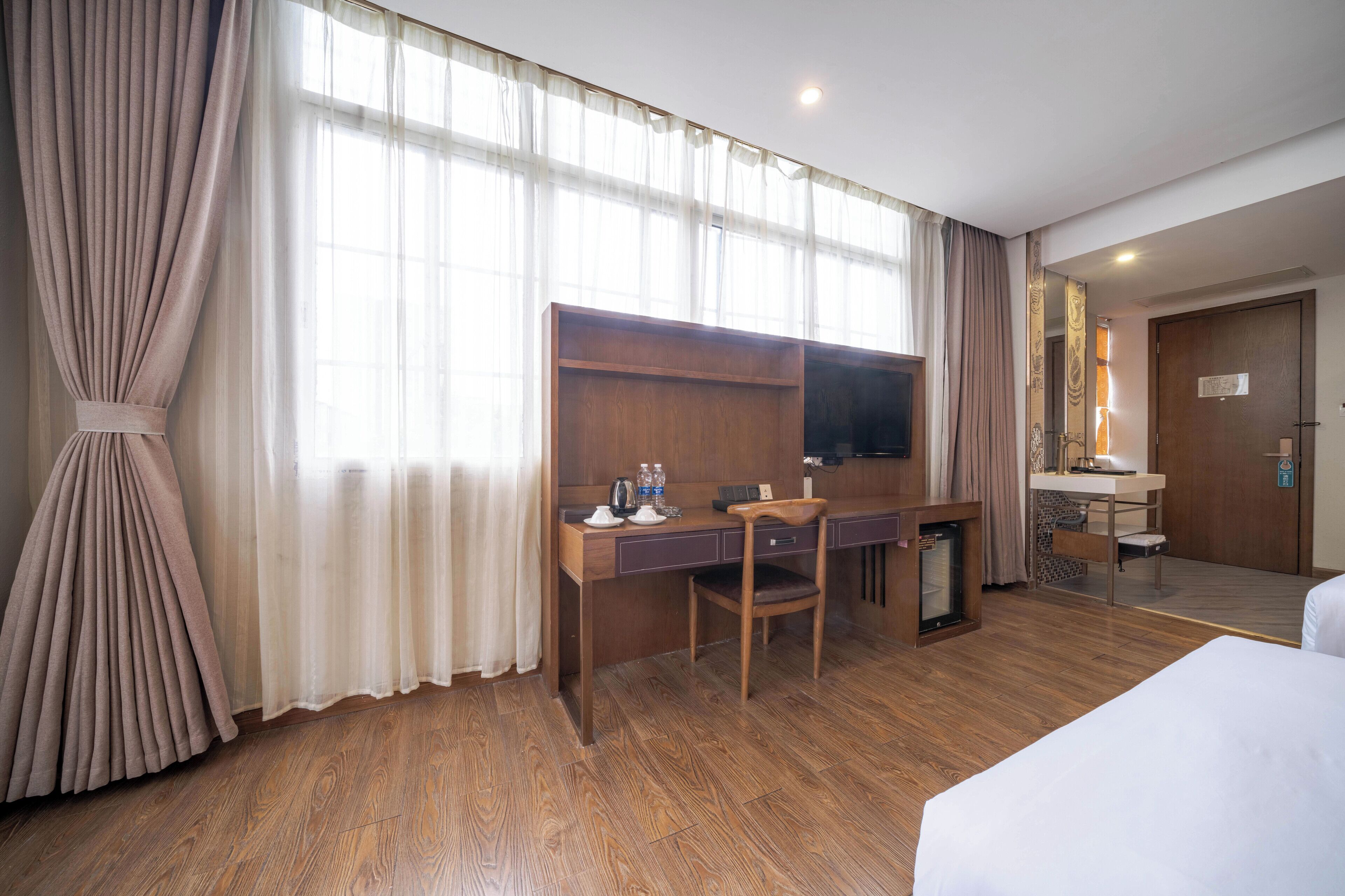 Ein Hotelzimmer mit Schreibtisch aus Holz, einem Sessel, einem Fernseher, einem Bett und einem großen Fenster mit Vorhängen.