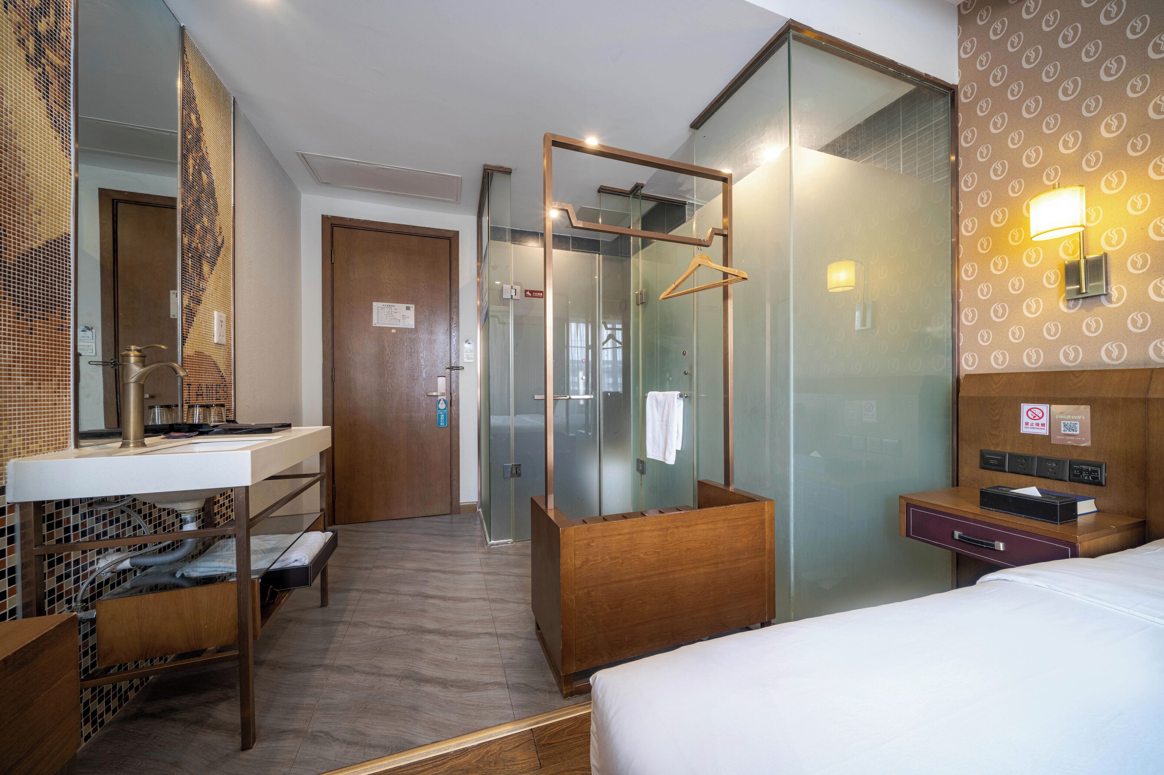 A hotel room with a bed, a wooden bedside table, a mirror, a glass shower enclosure, a wooden bench, and a wall-mounted light fixture.