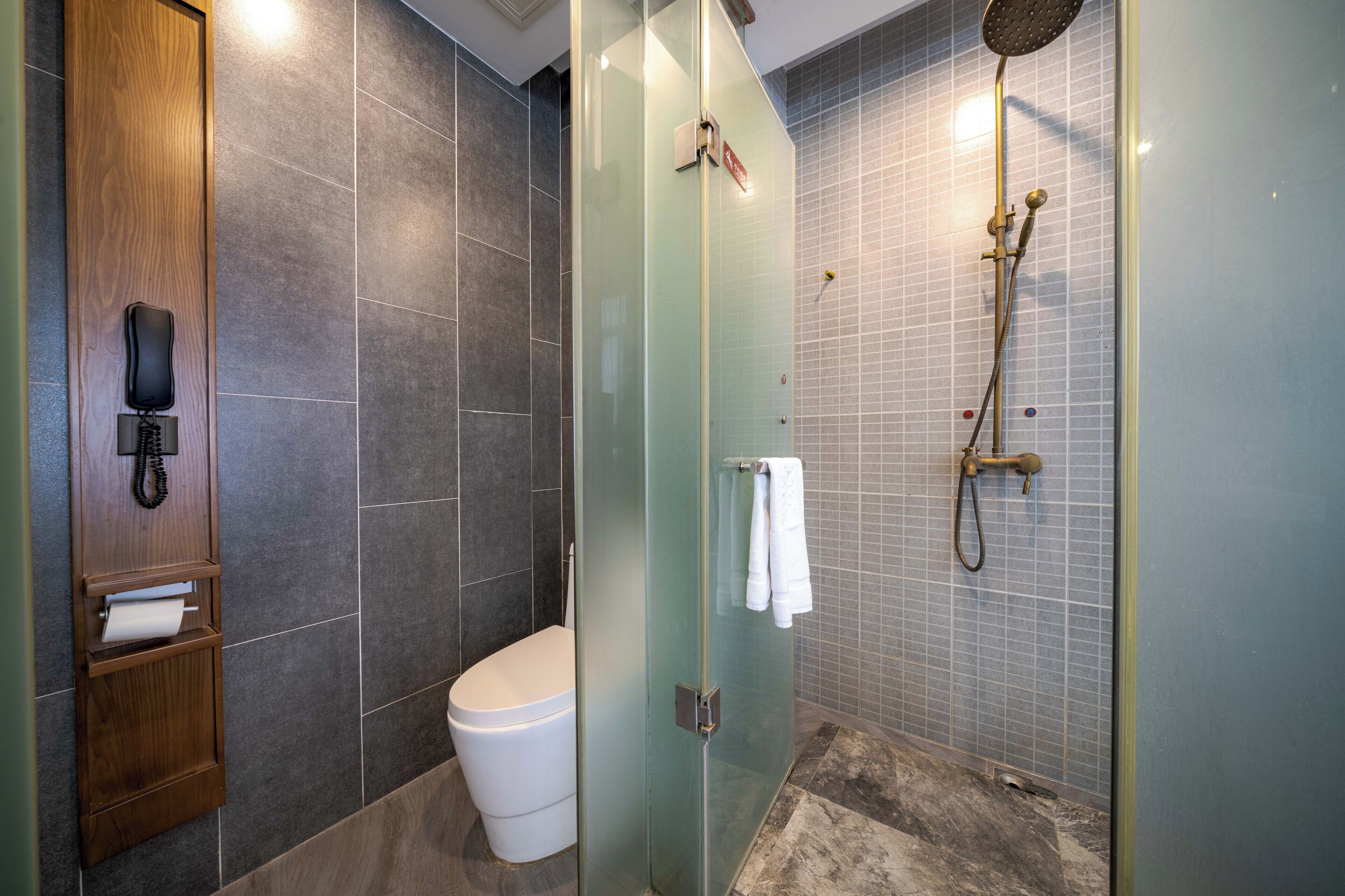 A modern bathroom with a glass-enclosed shower, a toilet, and a wooden telephone holder.