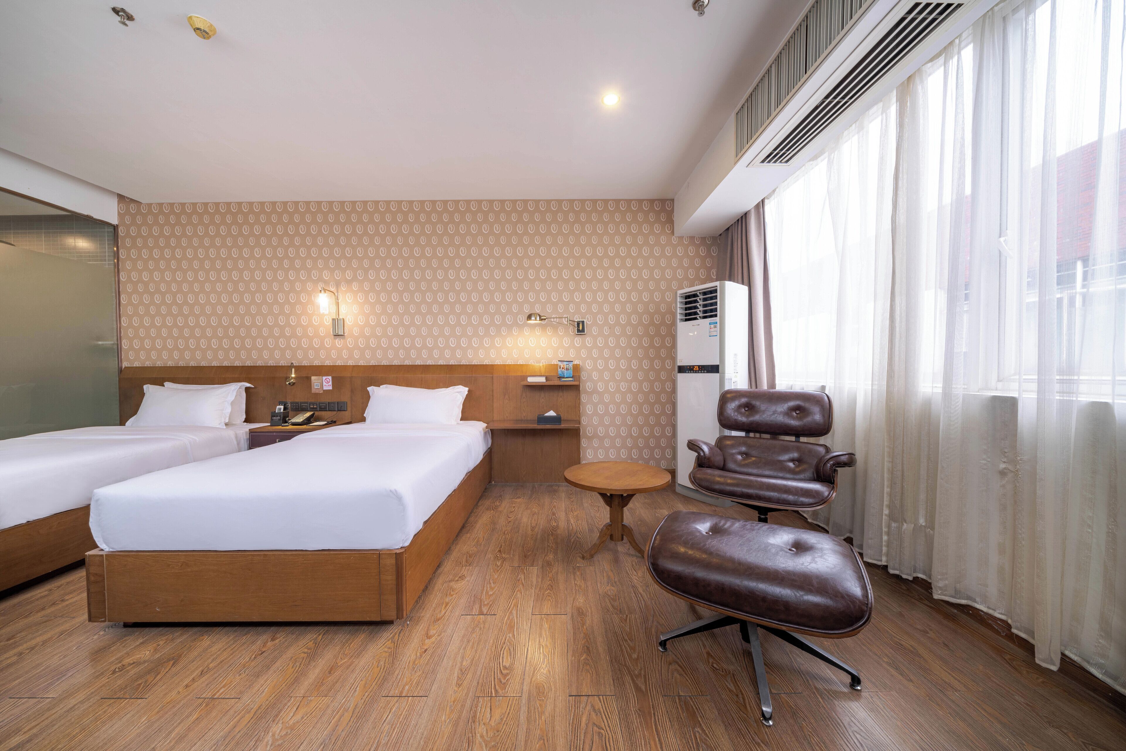 Ein Hotelzimmer mit zwei Betten, Holzboden, Ledersessel, kleinem runden Tisch und einem Fenster mit durchsichtigen Vorhängen.