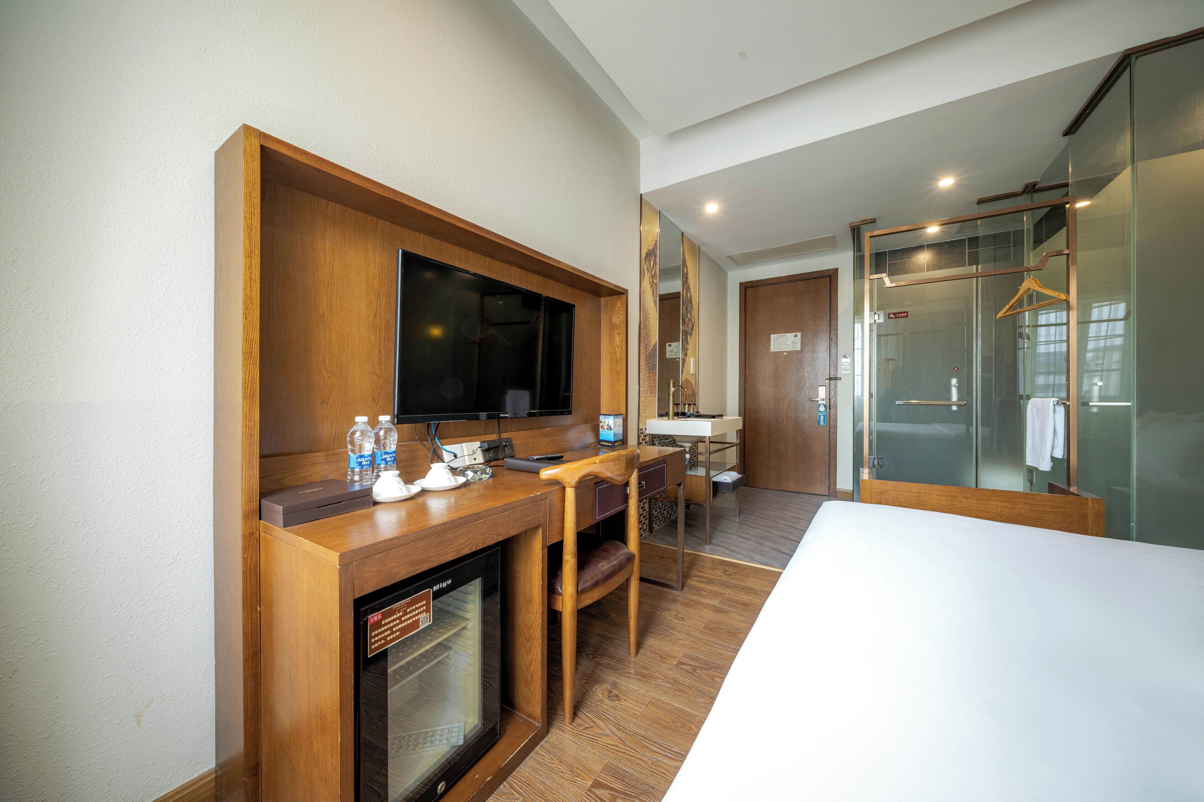 A hotel room with a wooden desk, a flat-screen TV, a minibar, a seating area with a chair and table, a large mirror, and a glass shower enclosure.