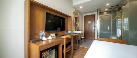 A hotel room with a wooden desk, a flat-screen TV, a minibar, a seating area with a chair and table, a large mirror, and a glass shower enclosure.