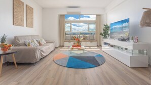 Een moderne woonkamer met een bank, salontafel en televisie. Er is een groot raam met uitzicht op de lucht en wolken.