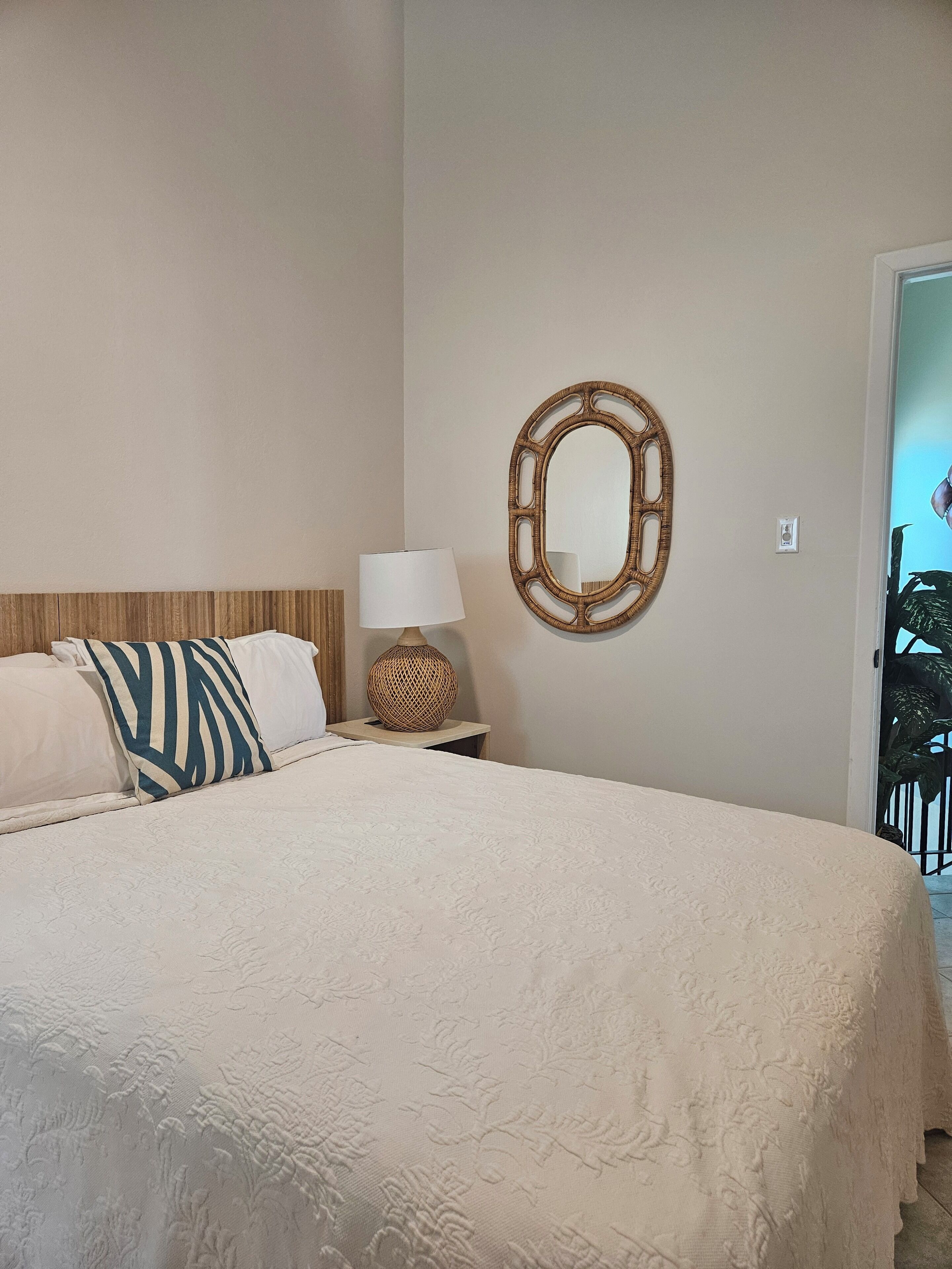 A neatly made bed with a white bedspread and two pillows, one with a striped pattern. A round mirror hangs on the wall above the bed, and a small table with a lamp and a woven basket is beside the bed.