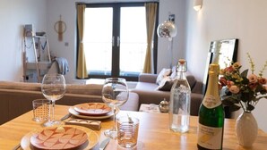 Ett matbord dukat med tallrikar, glas och en flaska mousserande vin, med en soffa och ett fönster i bakgrunden.
