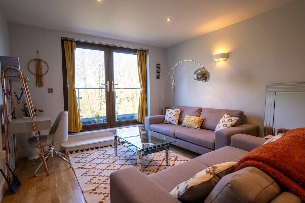 A modern living room with a sofa, a glass coffee table, and a desk with a chair. There is a large window with curtains, a lamp, and a radiator.