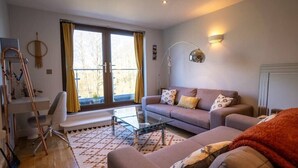 Ett modernt vardagsrum med en soffa, ett soffbord i glas och ett skrivbord med en stol. Det finns ett stort fönster med gardiner, en lampa och ett element.
