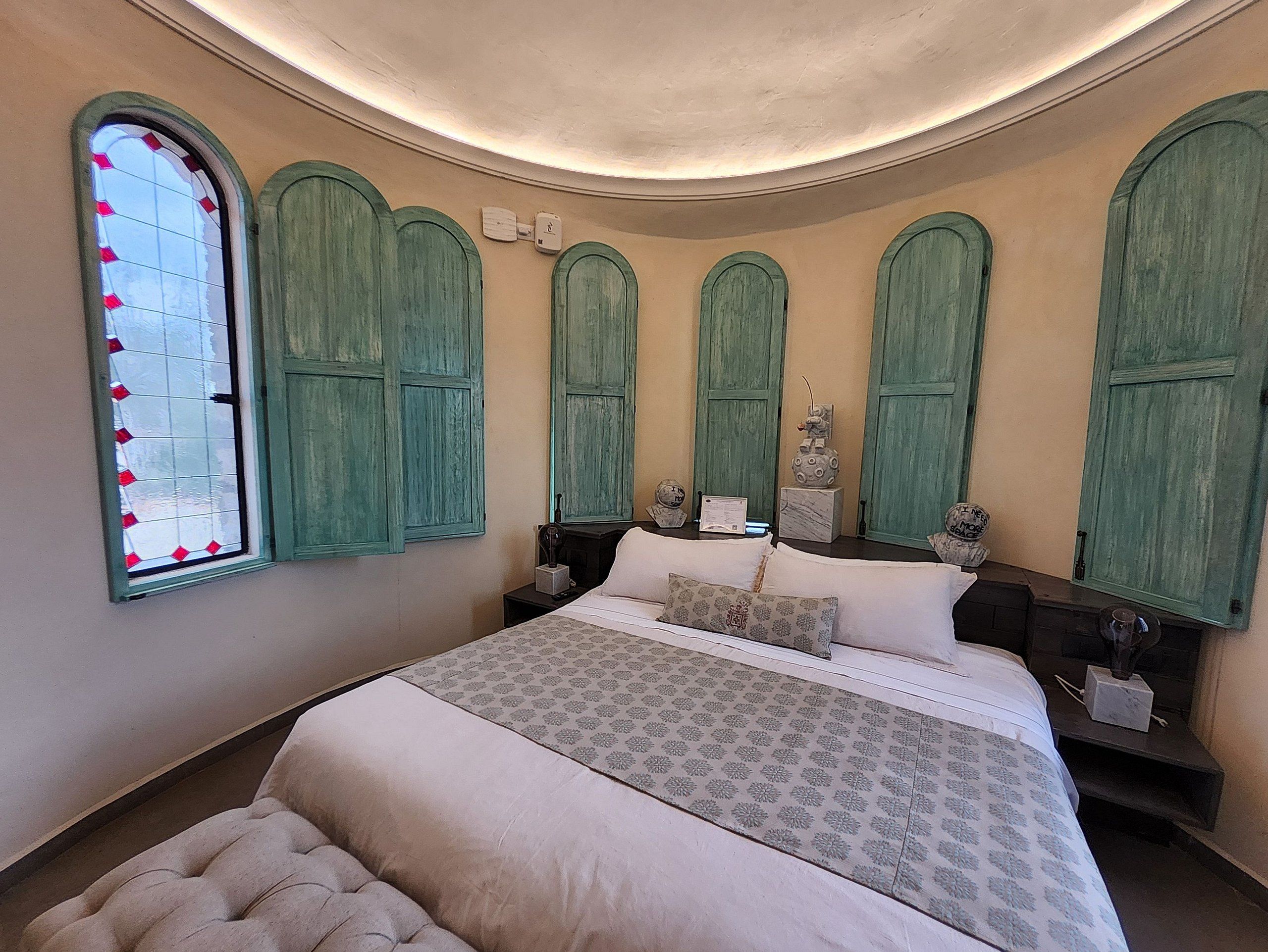 Ein Schlafzimmer mit einem großen Bett, grünen Rollläden, einem Zierfenster und einem kleinen Tisch mit einer Lampe.