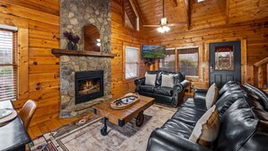 A log cabin interior with a stone fireplace, leather sofas, and a television. - Cozy 4BR Cabin 3 miles from Dollywood, Hot Tub, Game Room, Fast Wifi (Sevierville)