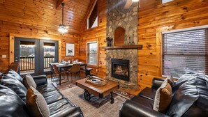A log cabin interior with a stone fireplace, leather sofas, a dining area, and a view through the door. - Cozy 4BR Cabin 3 miles from Dollywood, Hot Tub, Game Room, Fast Wifi (Sevierville)