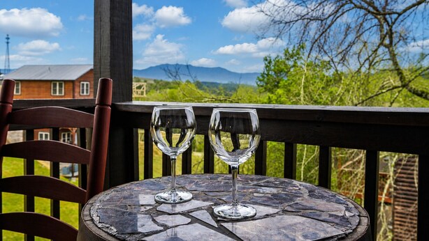 A table with two empty wine glasses on a stone-topped table, a wooden chair, and a view of a house and mountains in the background. - Cozy 4BR Cabin 3 miles from Dollywood, Hot Tub, Game Room, Fast Wifi (Sevierville)