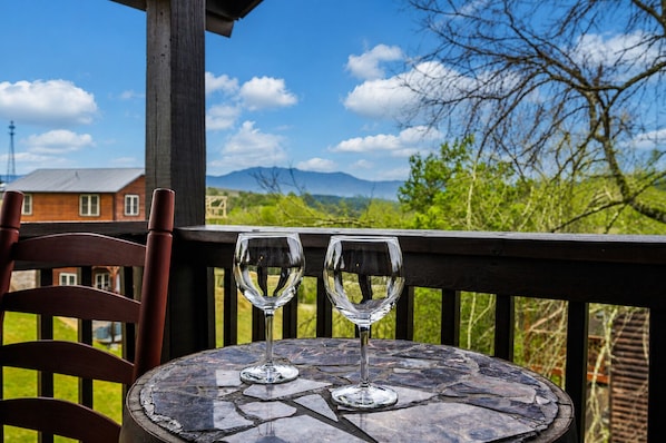 Une table avec deux verres à vin vides posés sur un plateau en pierre, une chaise en bois et, en arrière-plan, une vue sur une maison et des montagnes.