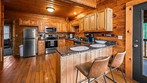 A wooden cabin-style kitchen with granite countertops, stainless steel appliances, and a bar area with stools. - Cozy 4BR Cabin 3 miles from Dollywood, Hot Tub, Game Room, Fast Wifi (Sevierville)
