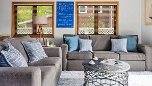 A living room with two grey sofas, a round coffee table, and a floor lamp. There are two windows with wooden blinds and a blue motivational poster on the wall. - All Sports Long Lake/Hot Tub/Private Beach/Dock/Kayaks/Gameroom/Pets OK (Traverse City)