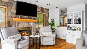 A living room with stone walls, a fireplace, two grey armchairs, a small round table, and a kitchen area with white cabinets and a coffee station. - All Sports Long Lake/Hot Tub/Private Beach/Dock/Kayaks/Gameroom/Pets OK (Traverse City)