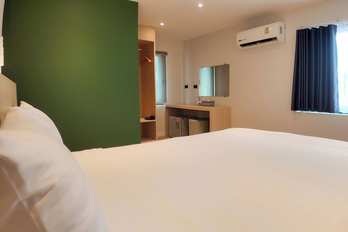 A hotel room with a bed, a desk with a mirror, an air conditioner, and a window with curtains.