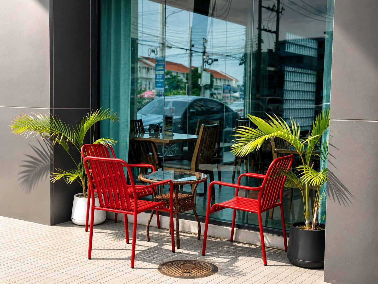 Un espacio al aire libre con sillas y mesas rojas, plantas en macetas y un escaparate de vidrio.