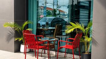Un espacio al aire libre con sillas y mesas rojas, plantas en macetas y un escaparate de vidrio.