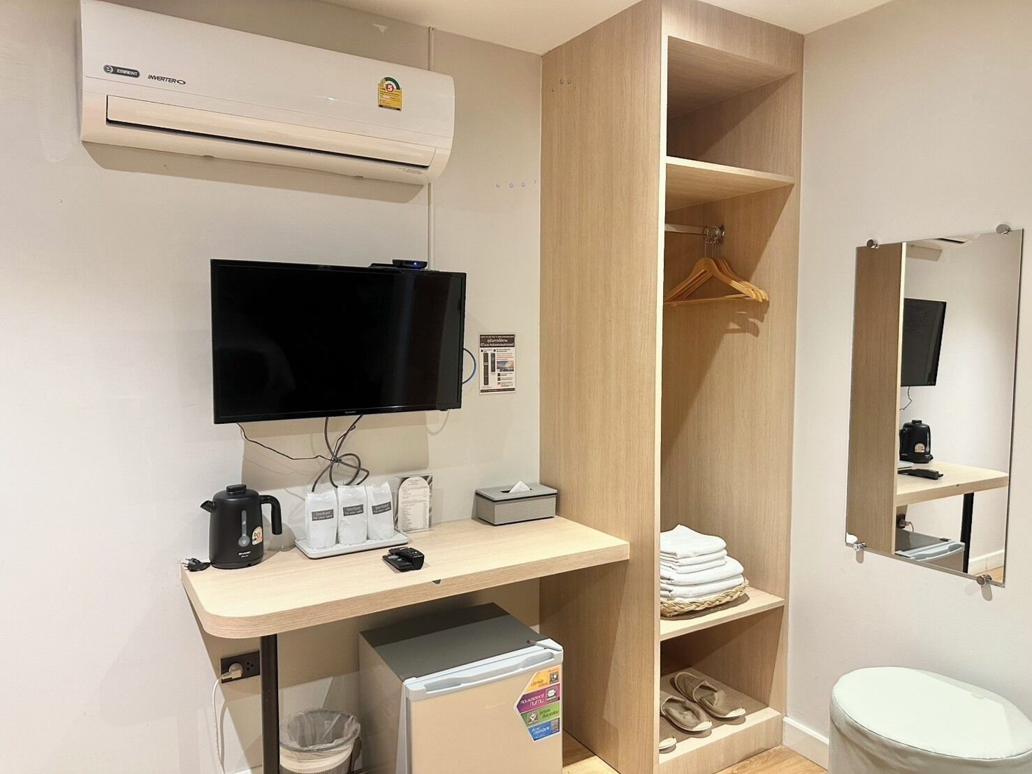Habitación de hotel compacta con televisor de pantalla plana, kitchenette pequeña, armario de madera, espejo y un inodoro.