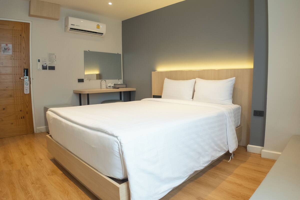 A modern hotel room with a large bed, a wooden desk, a mirror, and an air conditioning unit on the wall.