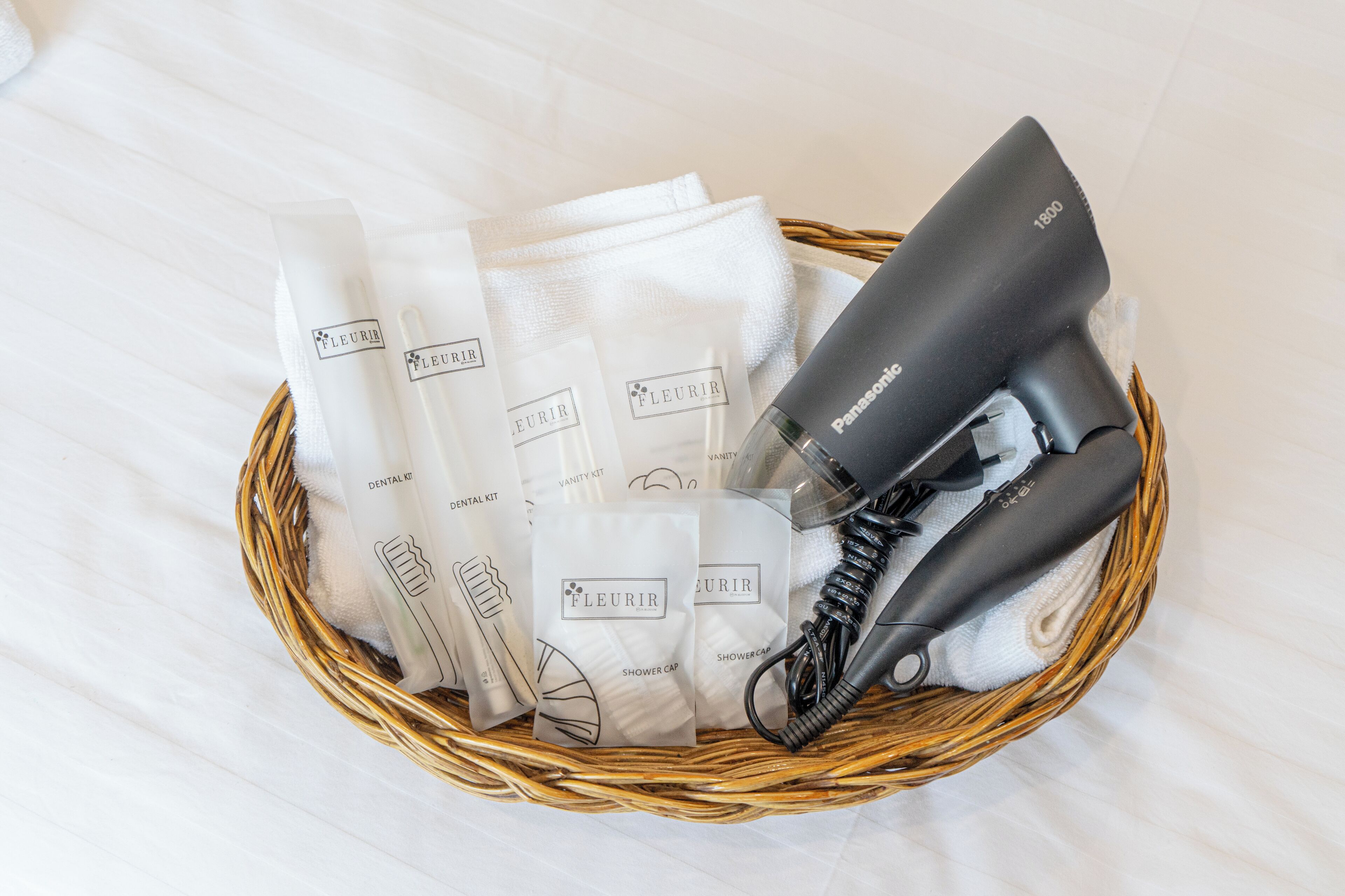 A wicker basket containing a Panasonic hair dryer, several white shower caps, and a dental kit.