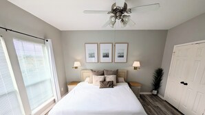 Une chambre à coucher avec un grand lit, deux lampes de chevet, un ventilateur de plafond et des tableaux encadrés accrochés au mur.
