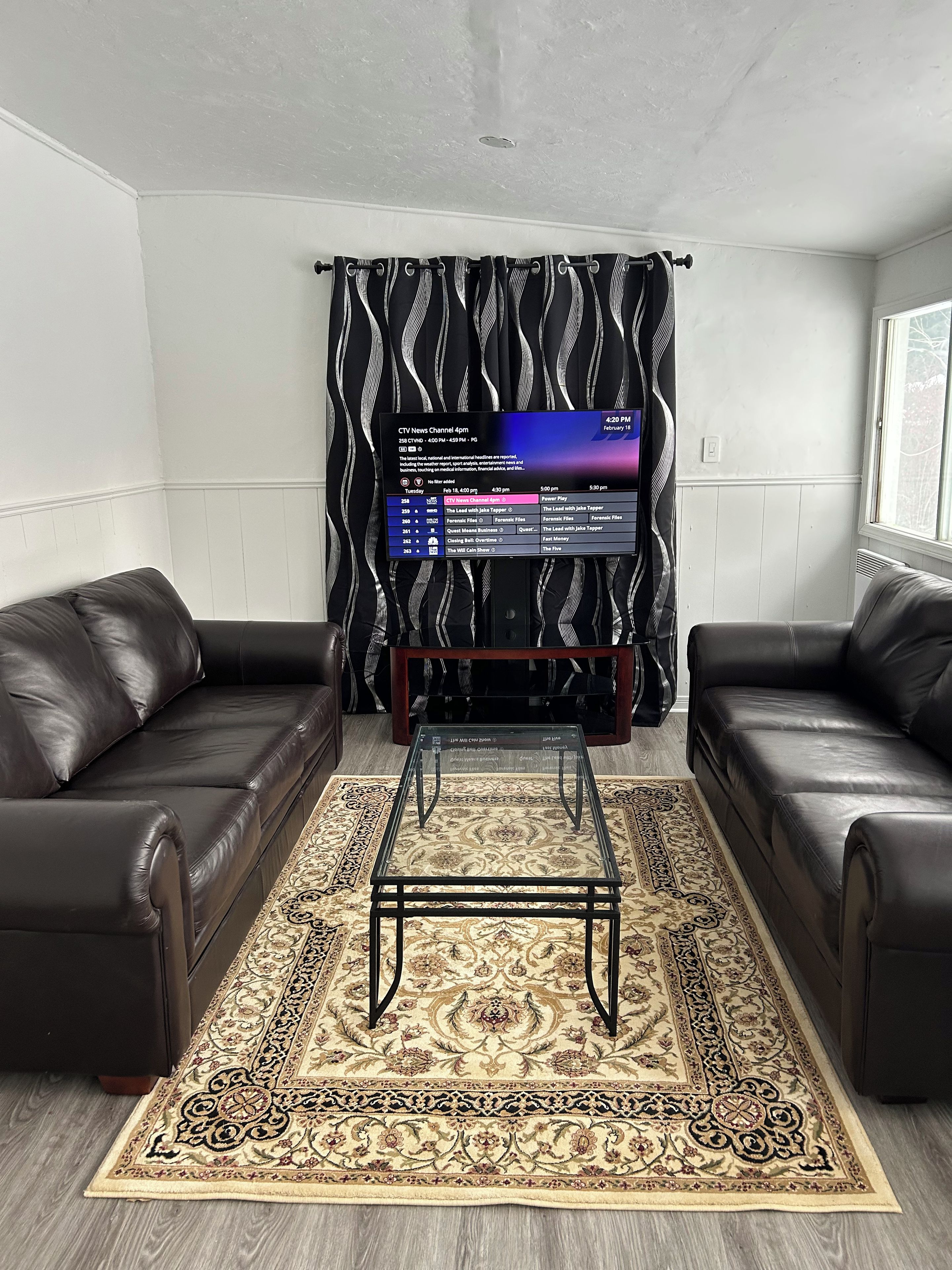 A living room with two black leather sofas, a black coffee table, a patterned rug, and a TV displaying a news channel schedule.