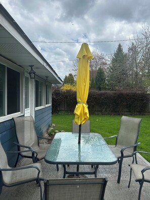 Une terrasse avec une table en verre et quatre chaises, un parasol jaune, et une maison en arrière-plan.