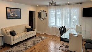 Una sala de estar moderna con un sofá color beige, una mesa de comedor de vidrio con sillas, un espejo grande y un cuadro de veleros.
