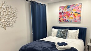 Un dormitorio con colcha azul marino, almohadas blancas y cabecera azul. Un cuadro abstracto y colorido está colgado en la pared sobre la cama.