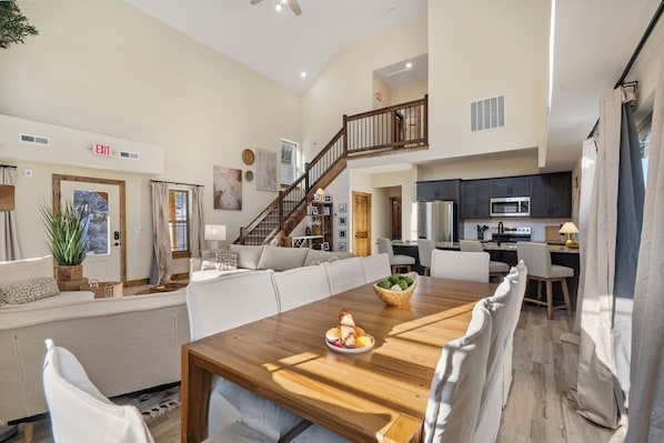 A spacious living area with a wooden dining table, a sofa, and a staircase leading to an upper level.