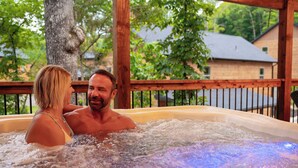 Una pareja en un jacuzzi con una terraza de madera y árboles al fondo.