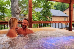 A couple in a hot tub with a wooden deck and trees in the background.